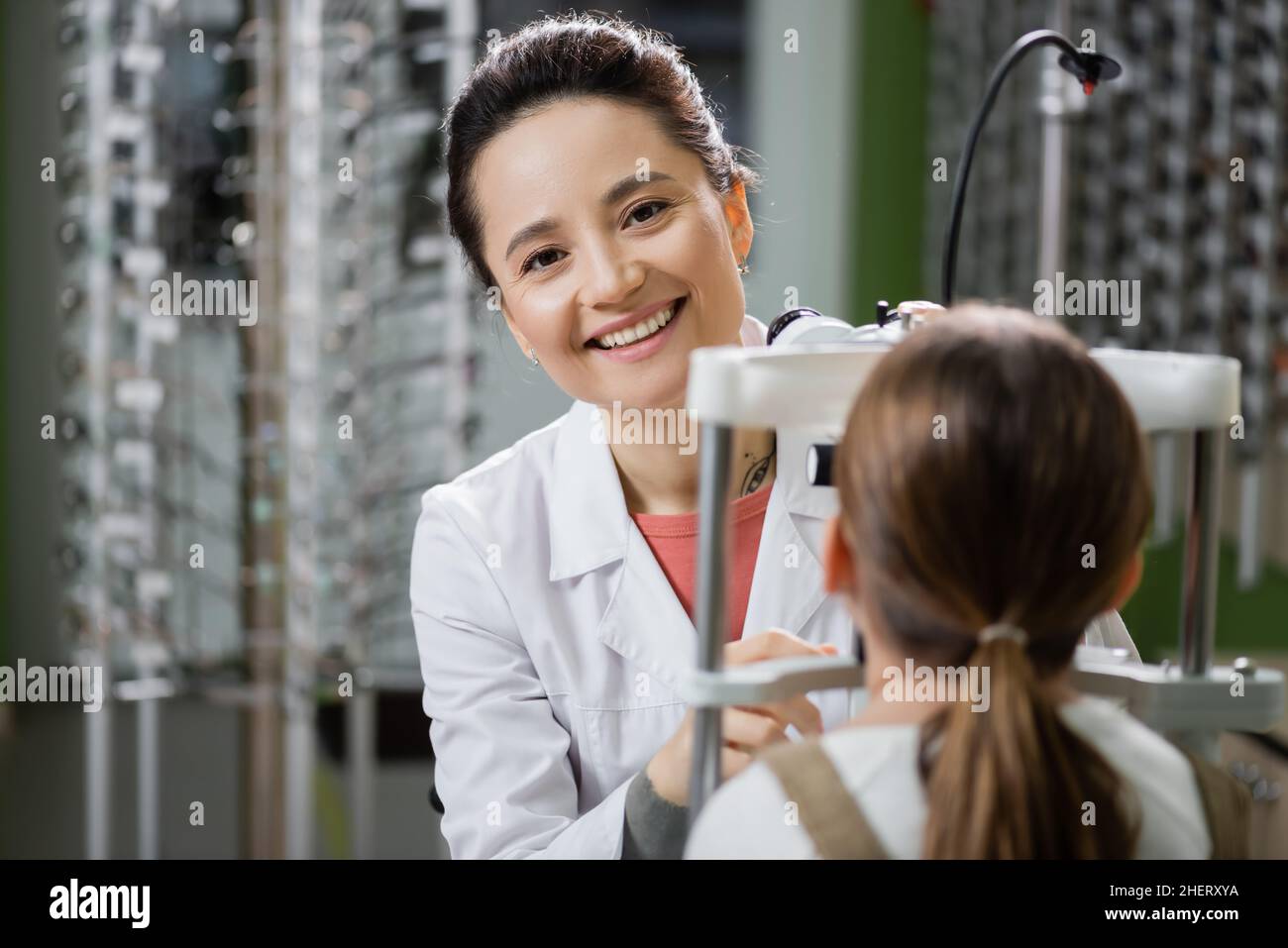 un oculista soddisfatto che guarda la macchina fotografica mentre controlla la vista della ragazza sul visore di visione in negozio di ottica Foto Stock