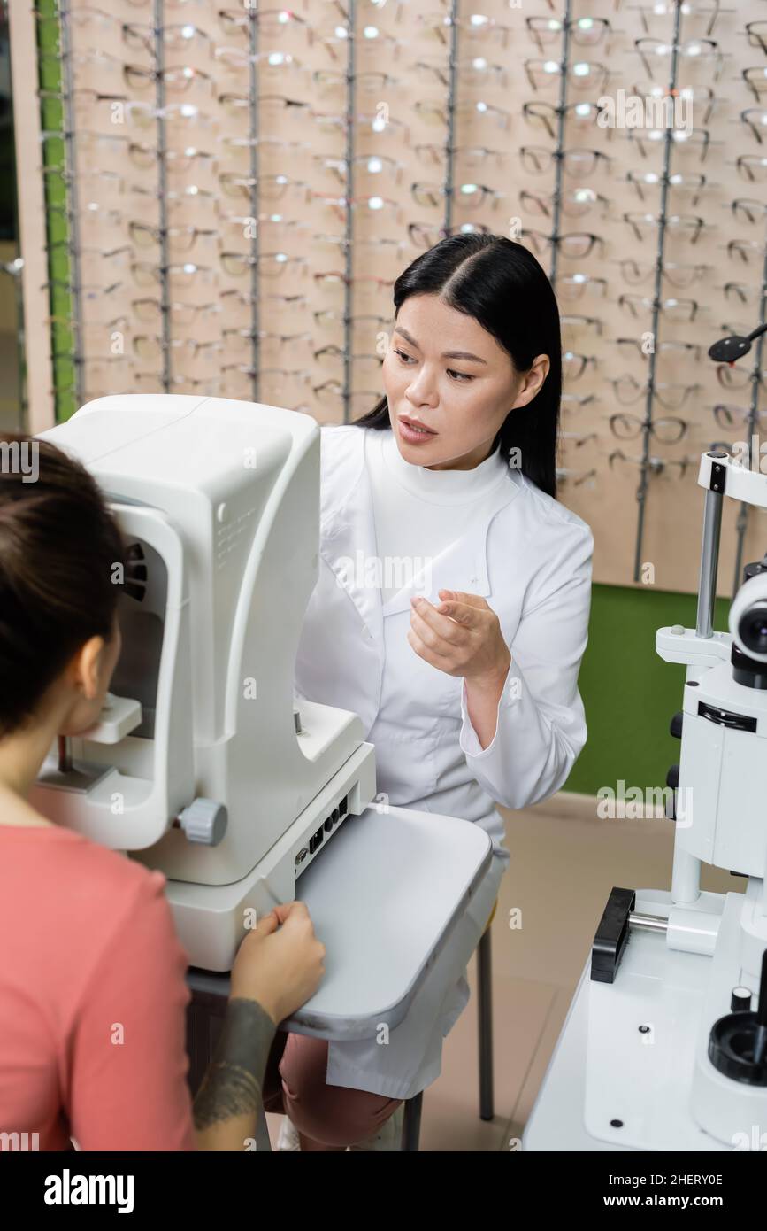 oftalmologo asiatico che punta con il dito mentre si misura la visione della donna sfocata su autorefrattore Foto Stock