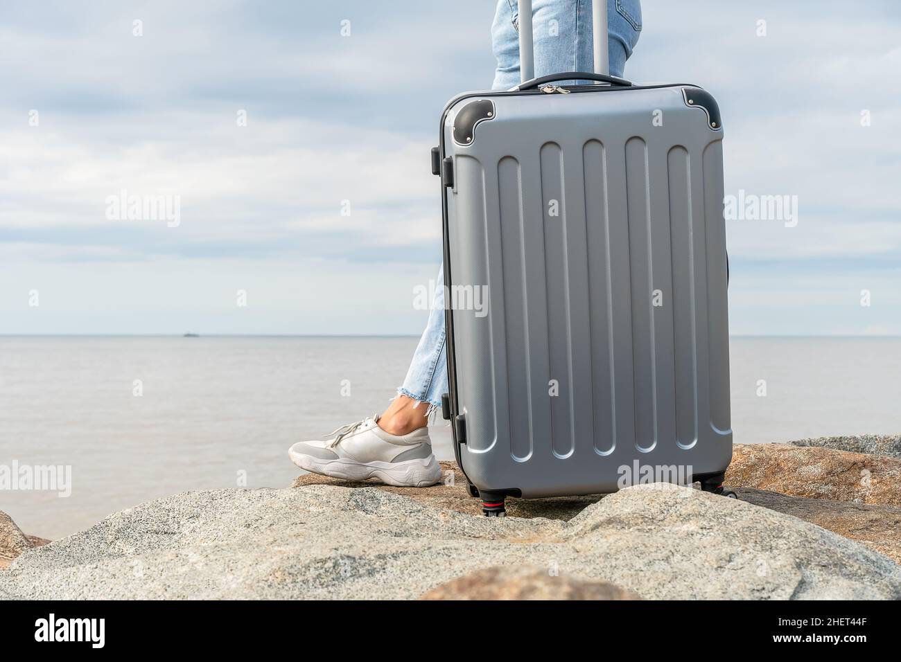 Primo piano di una valigia grigia e gambe di una donna in jeans sullo sfondo del mare e cielo con nuvole, spazio copia Foto Stock