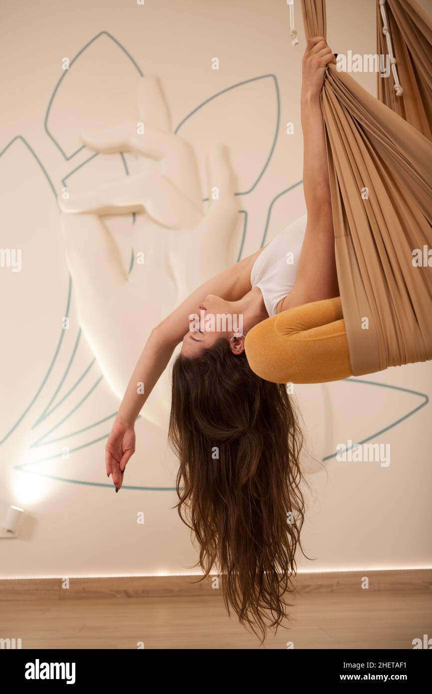 Scatto verticale corto di un gymnast femminile dai capelli lunghi che si allunga seduto in amaca da yoga aerea Foto Stock