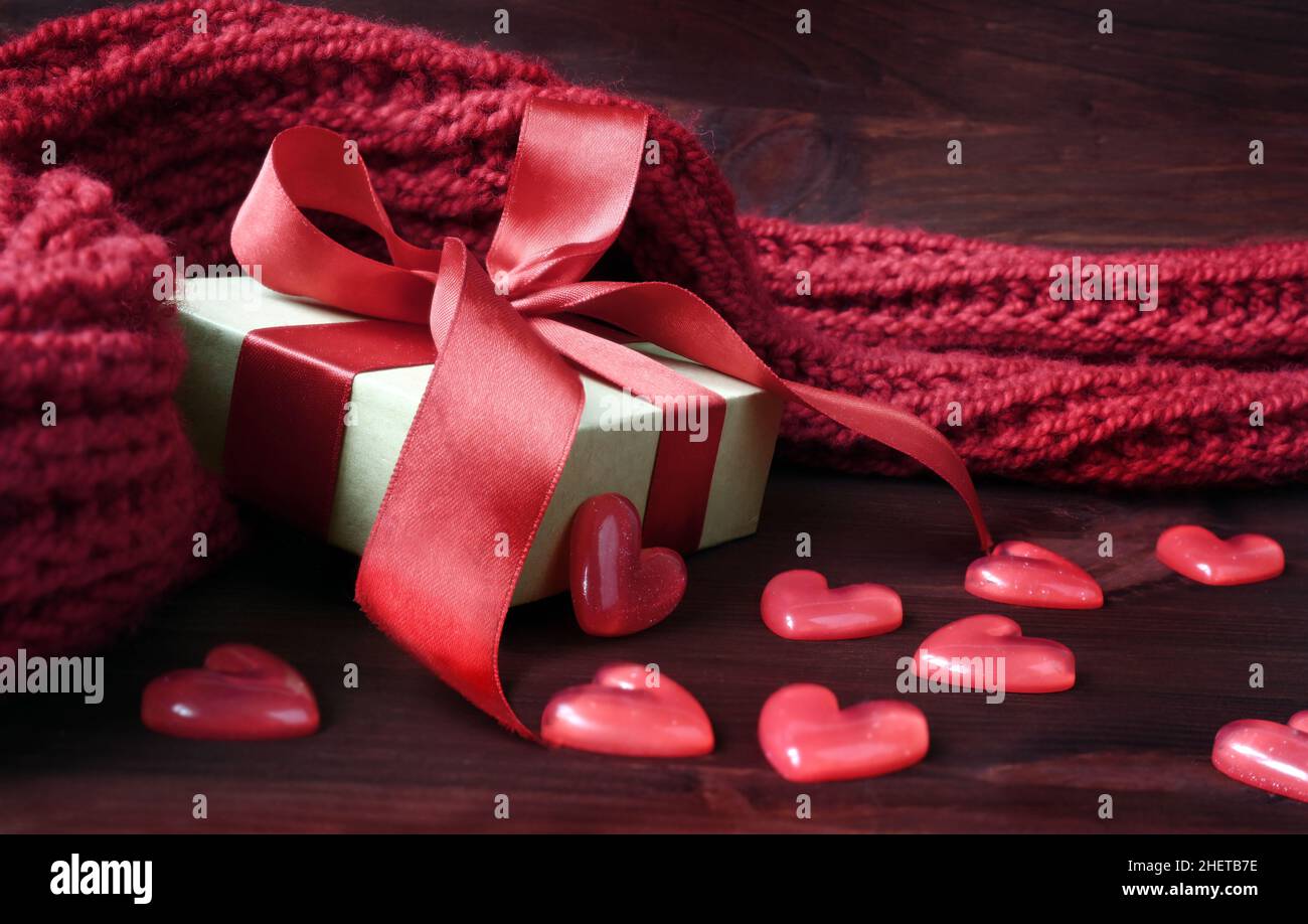 Confezione regalo bianca e cuori in plastica con sciarpa rossa in maglia su sfondo rosso in legno. Concetto di vacanza. Foto Stock