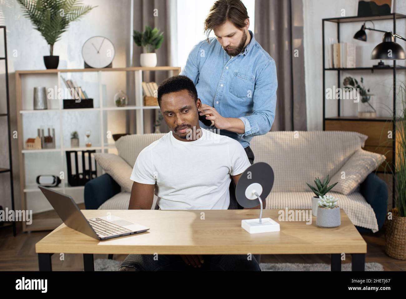 Vista frontale del bel maschio africano guardando nello specchio, mentre l'attraente ragazzo caucasico pulisce le setole rifilate dal collo dopo il taglio dei capelli Foto Stock