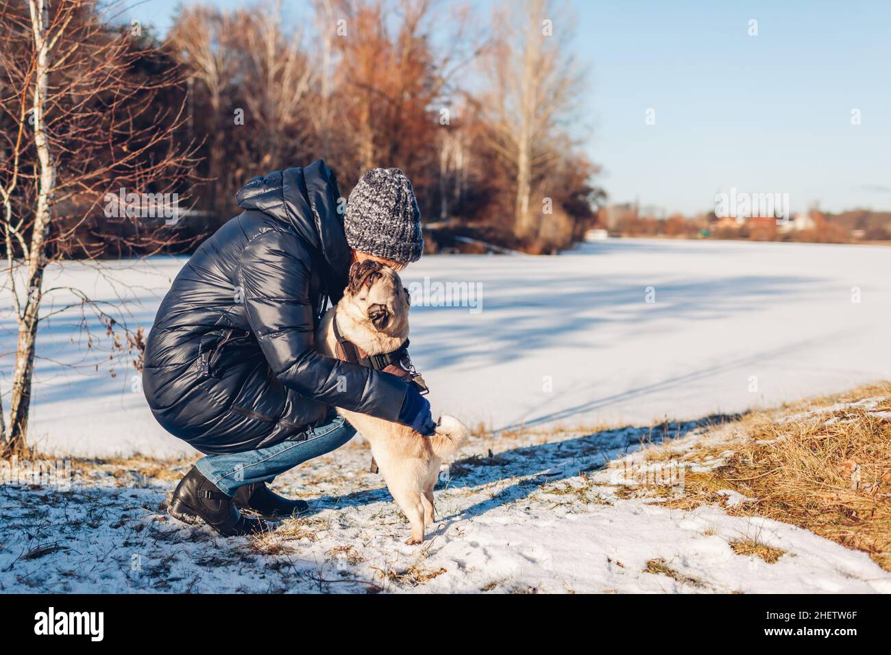 Donna che cammina pug cane nel parco nevoso invernale da lago ghiacciato abbraccio animale domestico. Buon cucciolo che indossa l'imbracatura in piedi sulle gambe posteriori. Foto Stock