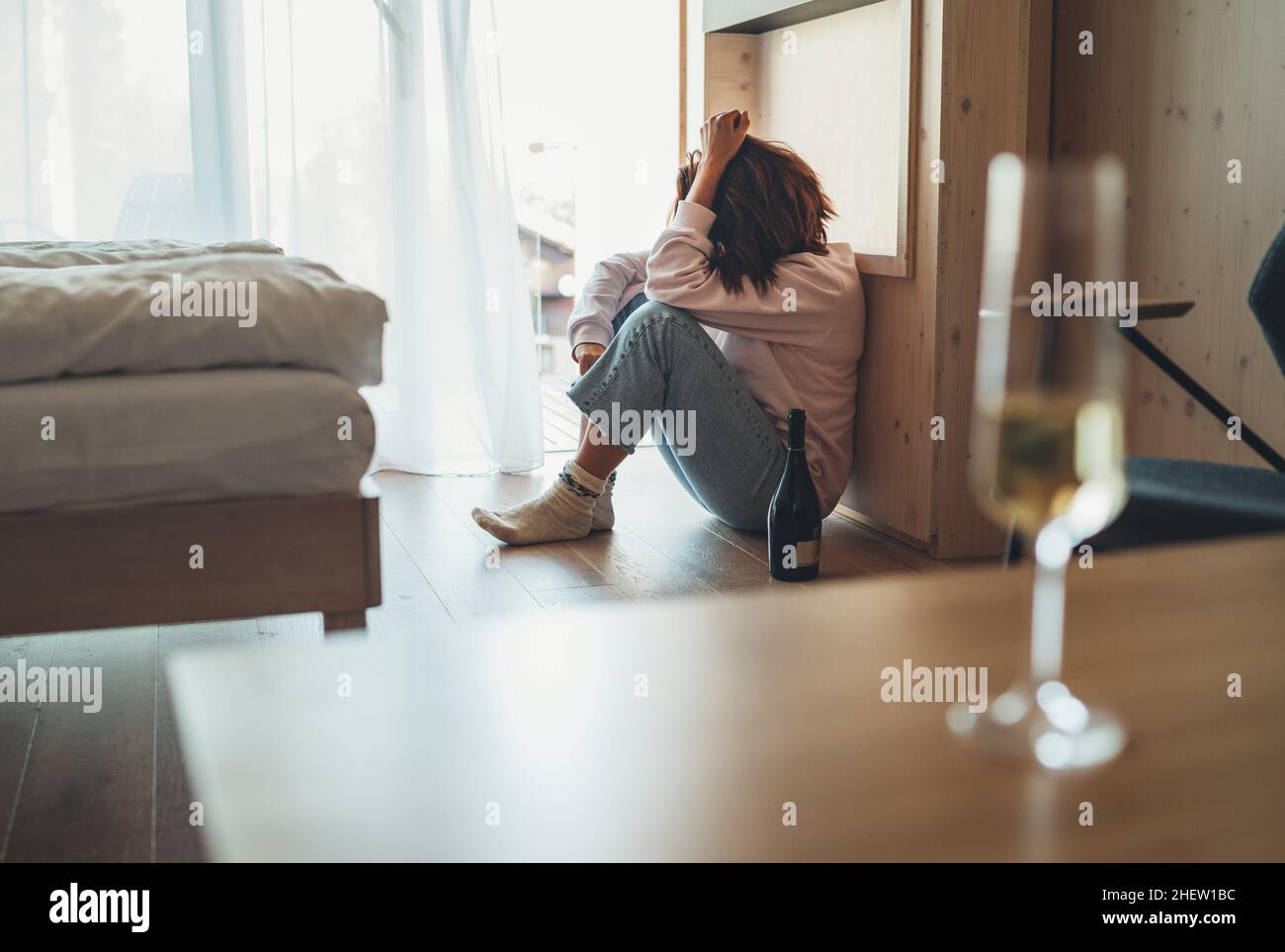 Donna triste seduta sul pavimento in camera da letto con una bottiglia di alcol. Bicchiere di vino non focalizzato sul tavolo in primo piano. Salute mentale e alcolismo pro Foto Stock