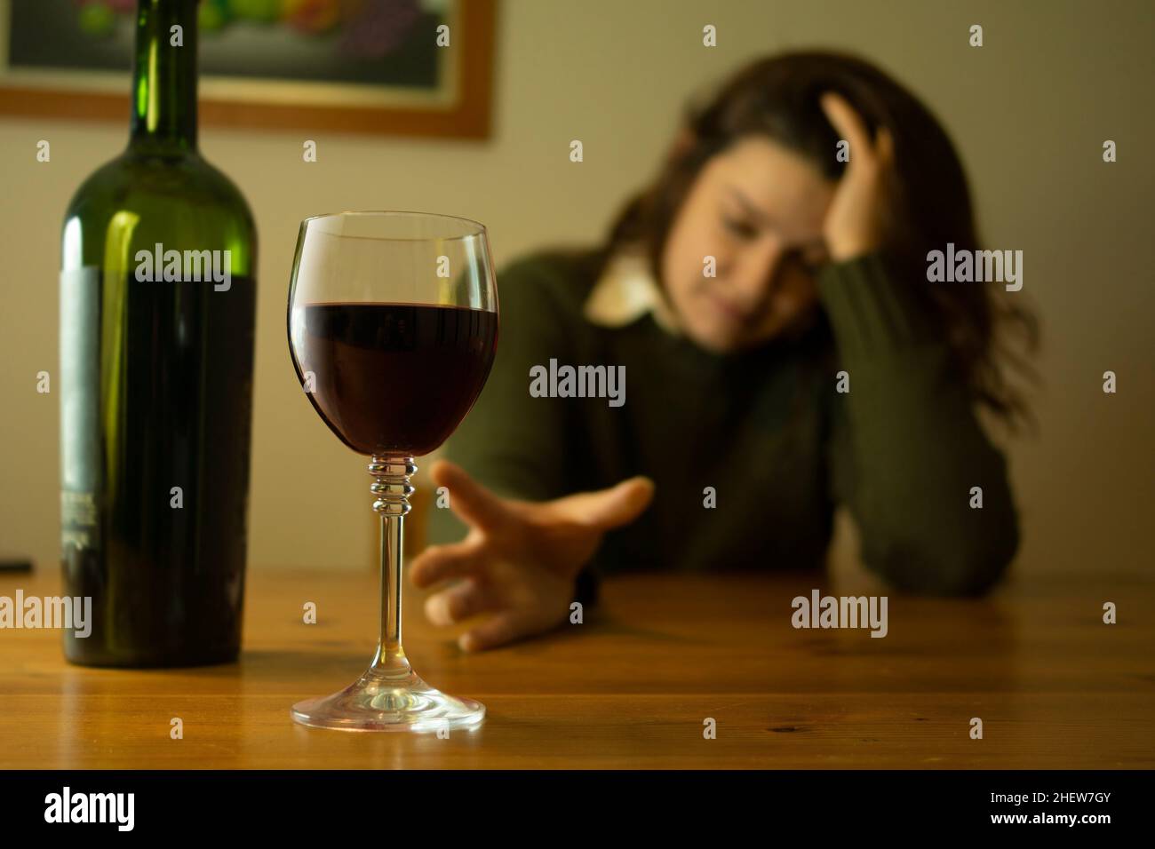 Primo piano su donna depressa con giacca verde seduta da sola ad un tavolo cercando di raggiungere un bicchiere di vino rosso con la mano Foto Stock