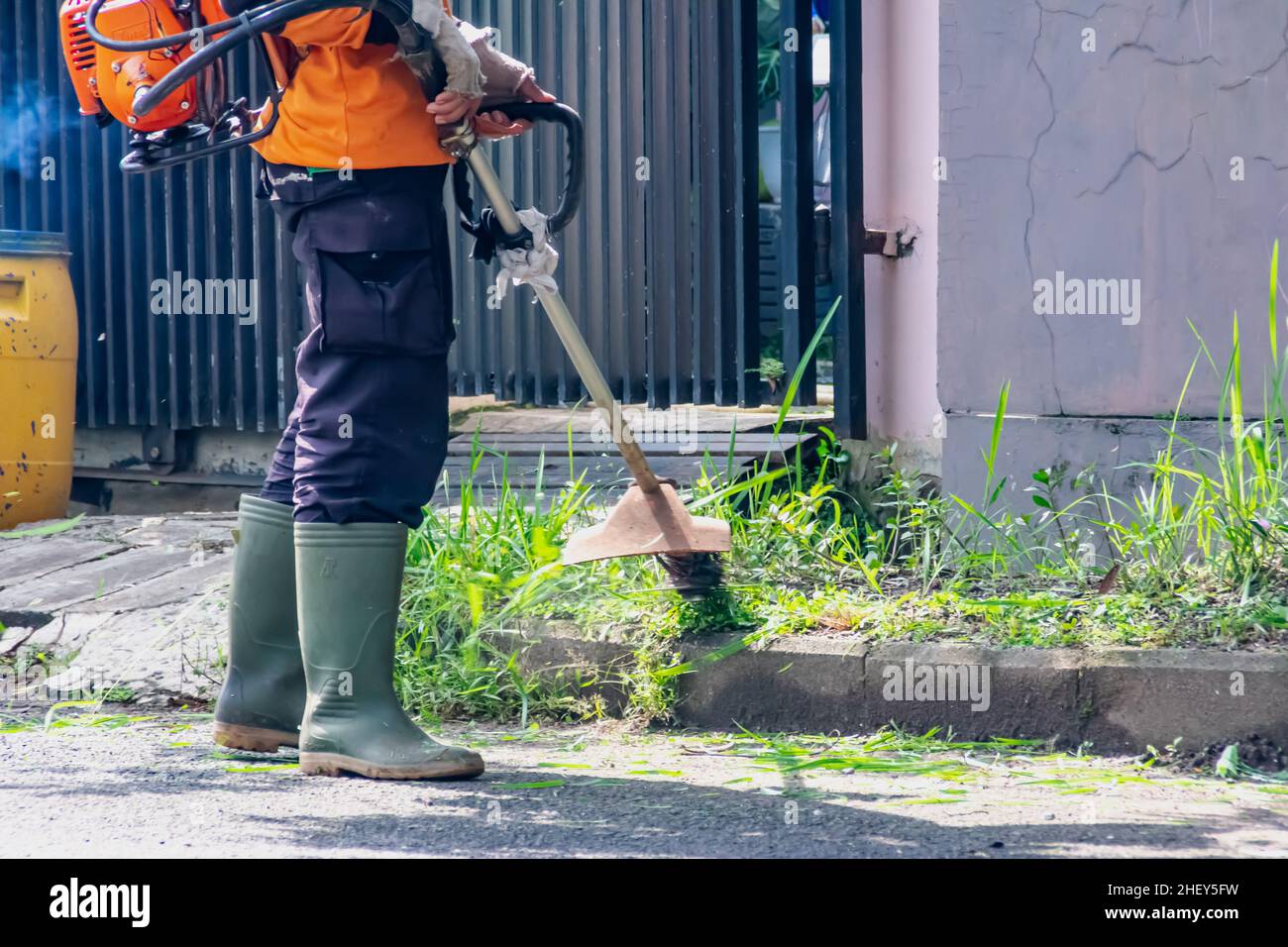 Uomo pubblico lavoratore tagliando l'erba del cortile della città con la macchina tagliatrice con un colpo di fumo via Foto Stock