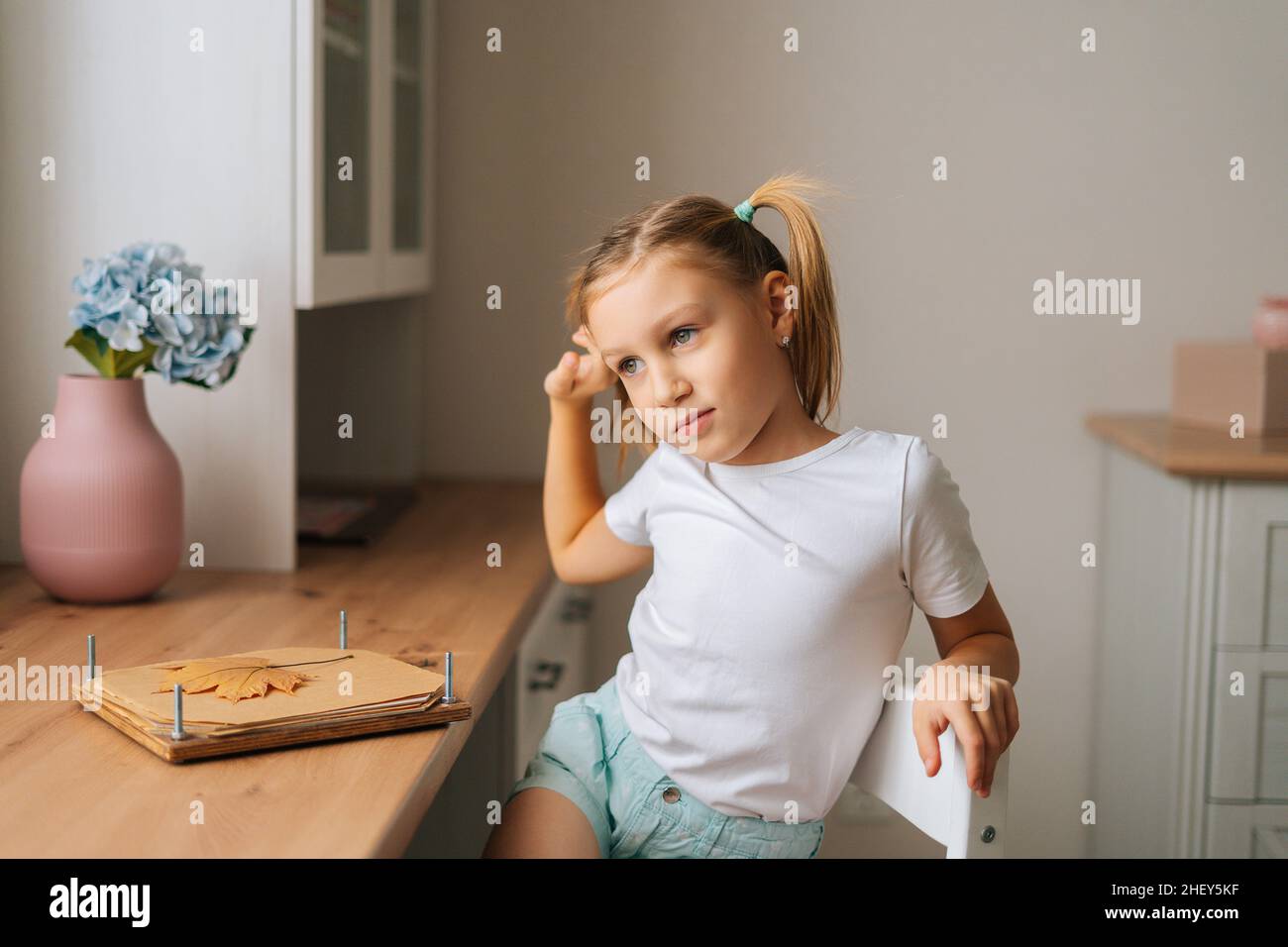 Ritratto della ragazza piccola primaria graziosa che si allunga fuori durante fare herbarium seduta al tavolo domestico dalla finestra. Foto Stock
