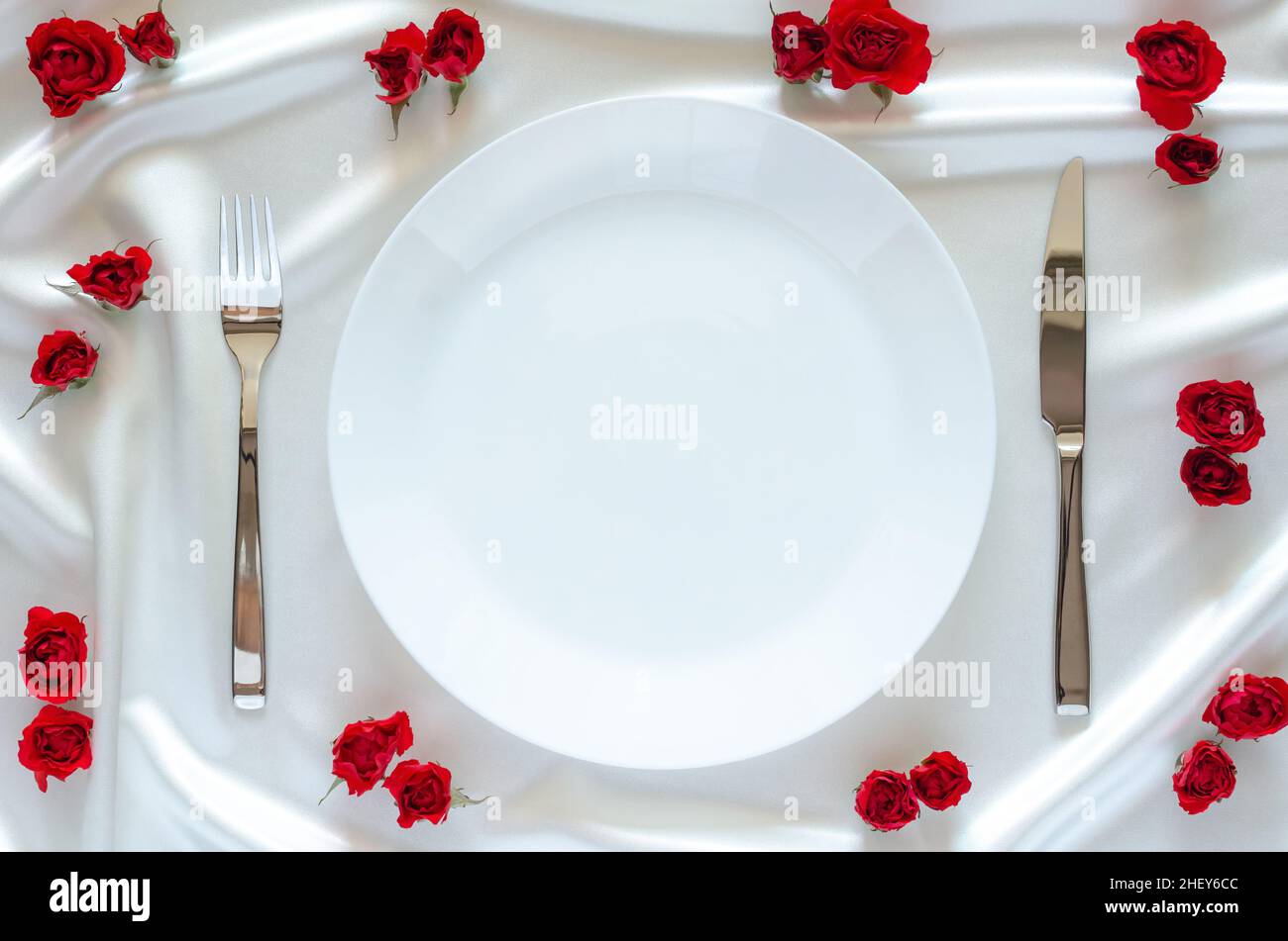 Piatto bianco con coltello e forchetta su tela satinata bianca e sfondo di rose rosse per cena, anniversario e San Valentino concetto. Foto Stock