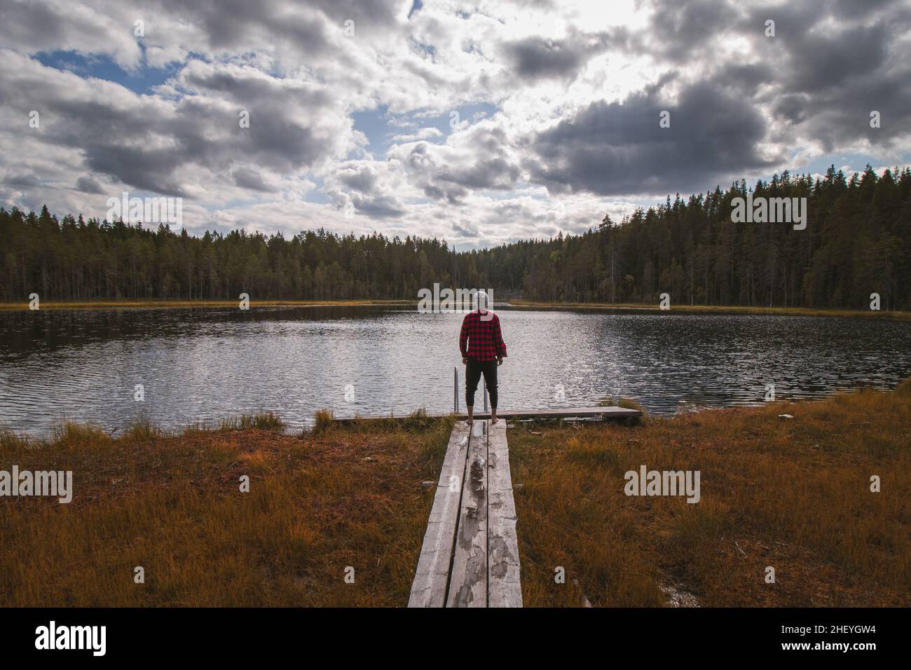 Esploratore e appassionato di viaggio in una camicia rossa e nera e berretto grigio si erge sul bordo di un passaggio in legno godendo la vista del lago Palolampi a Hii Foto Stock