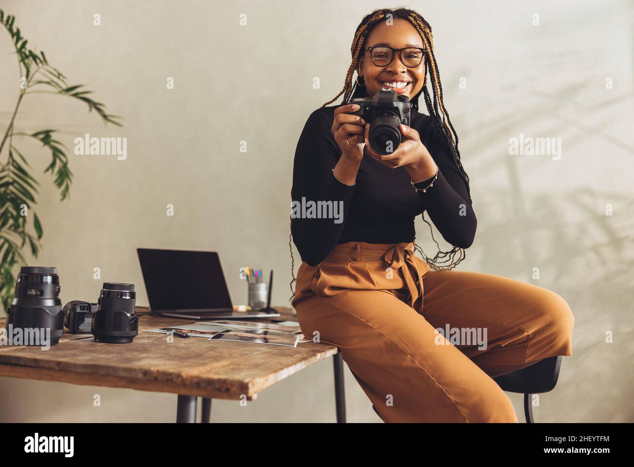 Fotografo sorridente alla fotocamera mentre si siede sulla scrivania. Felice giovane donna che tiene una fotocamera dslr nel suo ufficio domestico. Freelance artistico che lavora Foto Stock