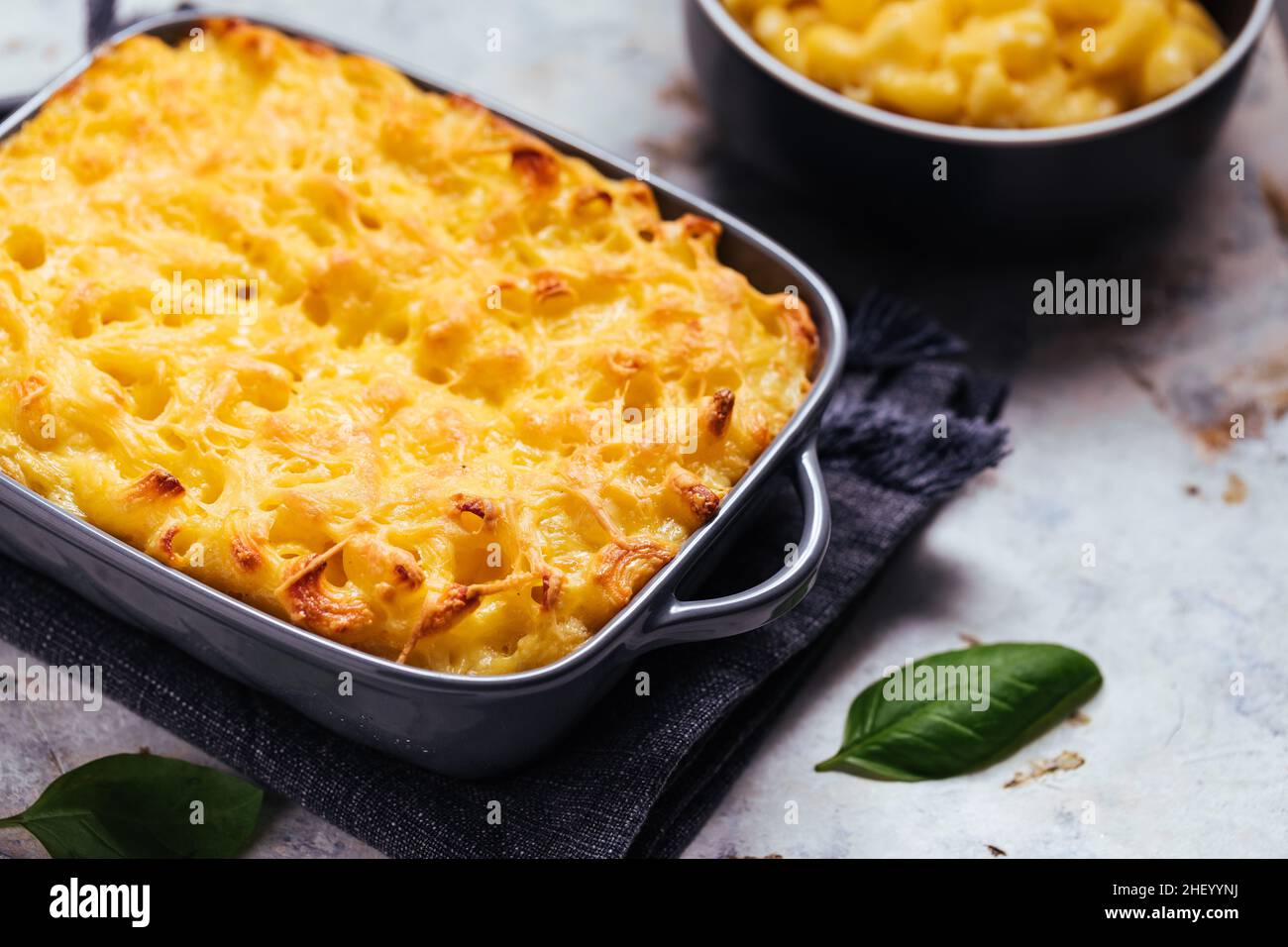 mac classico e formaggio con basilico sulla parte superiore e mac e formaggio cotto in forno sullo sfondo con forchette e macaroni crudi su sfondo rusticžwooden Foto Stock