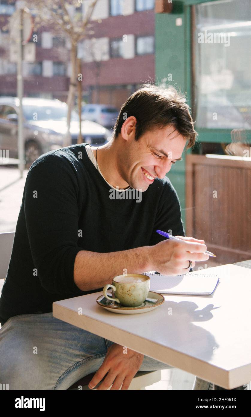 giovane uomo in una giacca grigia che scrive qualcosa nel libro mentre prende un caffè al bar Foto Stock