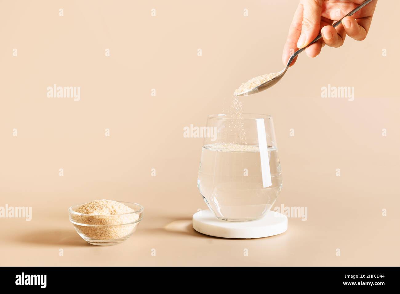 Donna aggiunge fibra di psyllium al vetro di acqua su sfondo beige. Superfood per intestino sano e dieta senza glutine. Foto Stock
