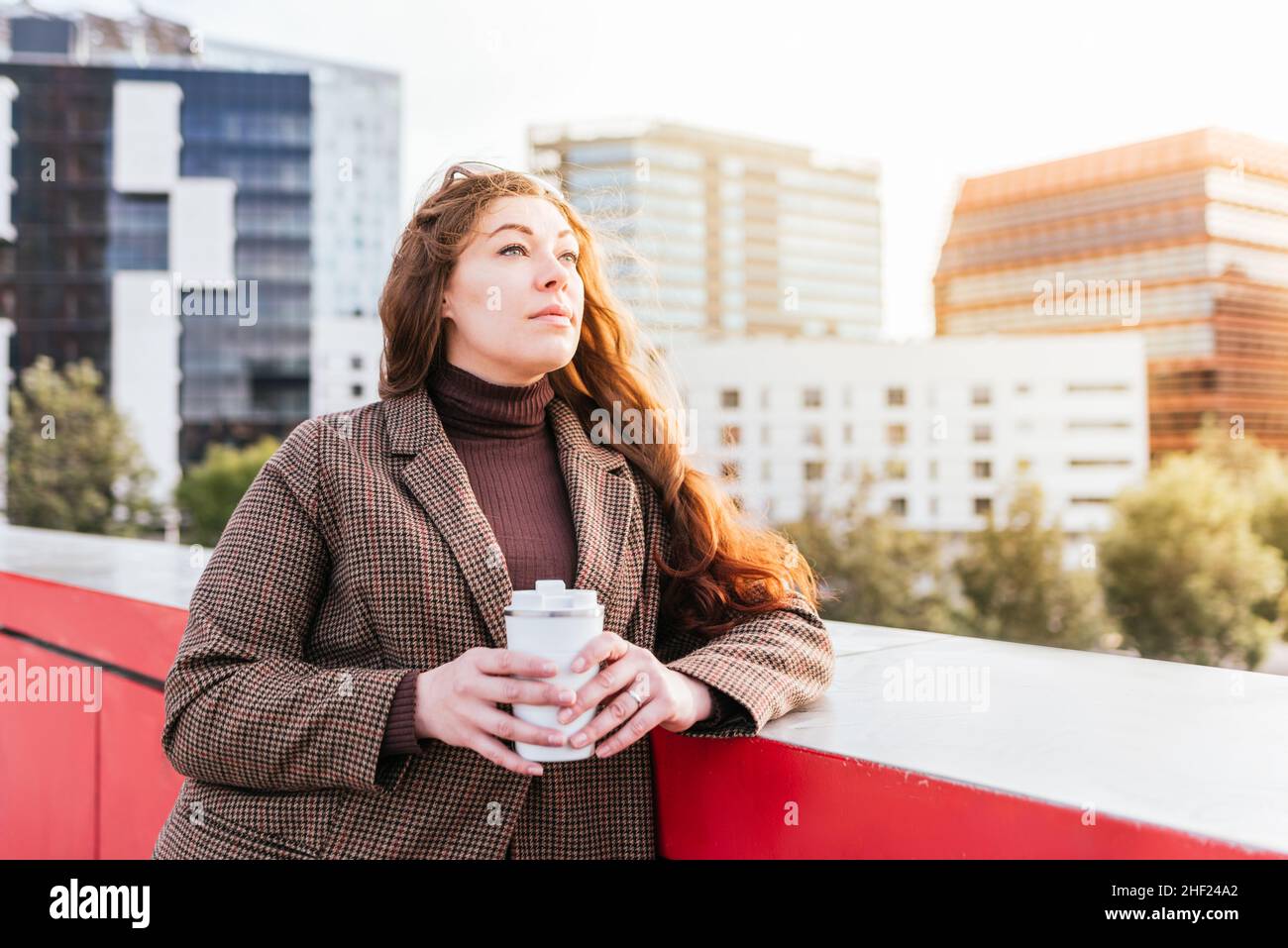 Donna pensiva in outerwear con capelli lunghi. Sta guardando via e pensando mentre si appoggia sul bordo del ponte e godendo il caffè da asporto in giorno di sole in città Foto Stock