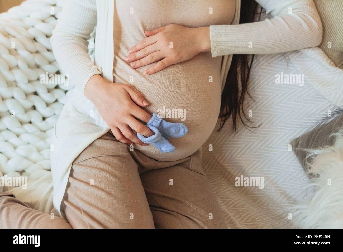 Concetto di maternità e gravidanza. Foto ritagliata di donna incinta che tiene un paio di calze carino blu per il bambino non nato sul ventre mentre si rilassa a casa Foto Stock