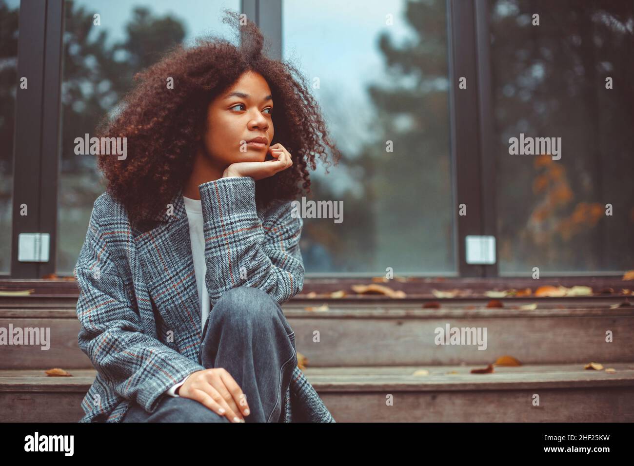 Ritratto all'aperto autunnale di giovane donna afroamericana pensierosa guardando da parte seduta su scale di casa in legno vecchio, ragazza curly pensiva passare il tempo Foto Stock