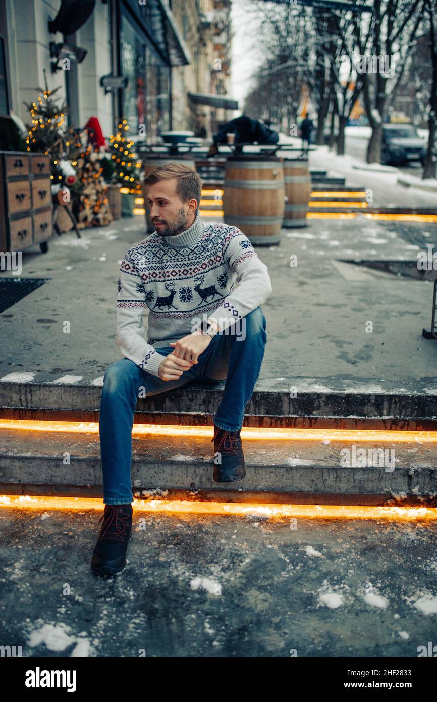 Il giovane uomo si siede sulle scale e guarda con cura alla strada decorata per Natale. Foto Stock