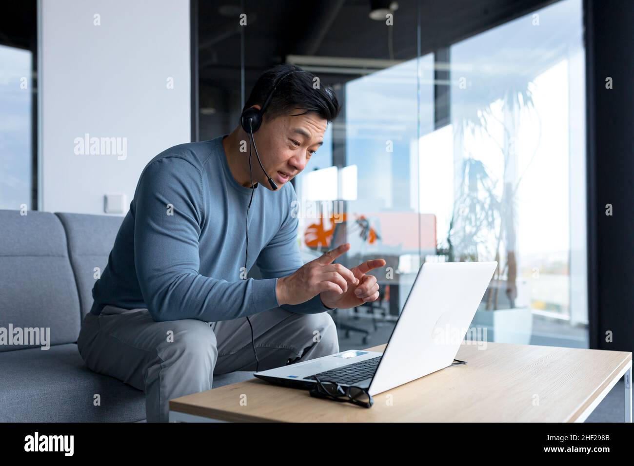 Un uomo asiatico felice e sorridente in un ufficio moderno utilizza una cuffia per una videochiamata, un uomo d'affari sorride all'interlocutore e guarda la paura del computer portatile Foto Stock