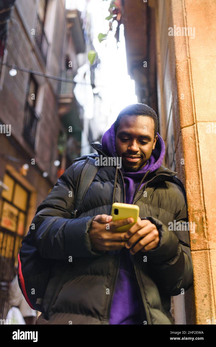 Basso angolo di uomo afroamericano in casual outerwear con zaino. Sta appoggiandosi sulla parete della costruzione e sta esplorando i social media sul telefono mobile sulla strada della città Foto Stock