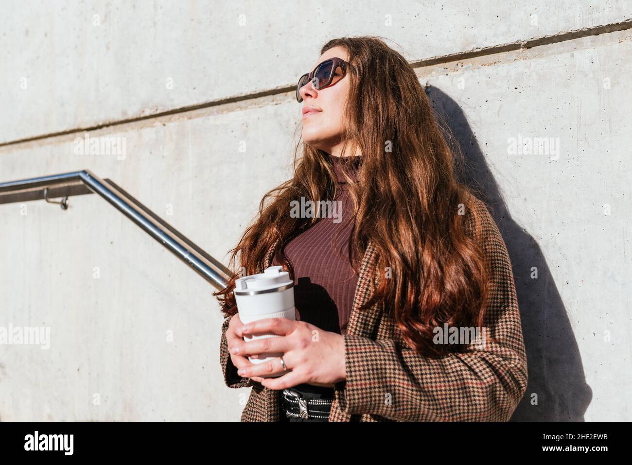 Angolo di donna basso con tazza di caffè ecologico da asporto appoggiata su pareti in cemento e guardando via nella giornata di sole in città Foto Stock