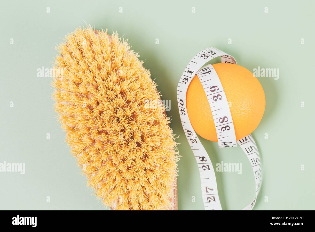 Spazzola per massaggio anticellulite accanto all'arancione e nastro di misurazione su sfondo verde, primo piano, vista dall'alto Foto Stock
