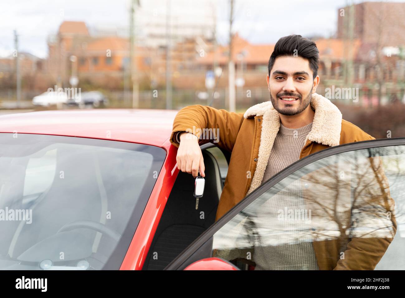 Uomo latino o arabo vicino all'auto che tiene le chiavi con la porta aperta sul parcheggio della città Foto Stock
