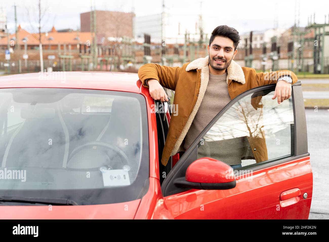 Uomo latino o arabo vicino all'auto che tiene le chiavi con la porta aperta sul parcheggio della città Foto Stock