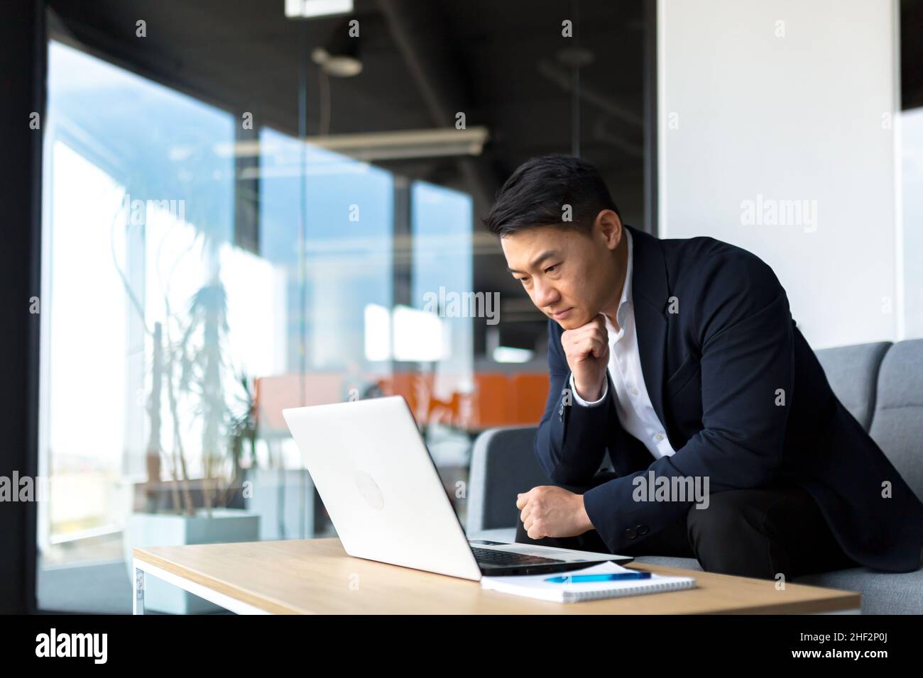 Un uomo d'affari asiatico attento al ritratto che lavora su un computer portatile in una moderna scrivania da ufficio. Uomo sicuro e concentrato in tuta formale in interni. Sottile Foto Stock