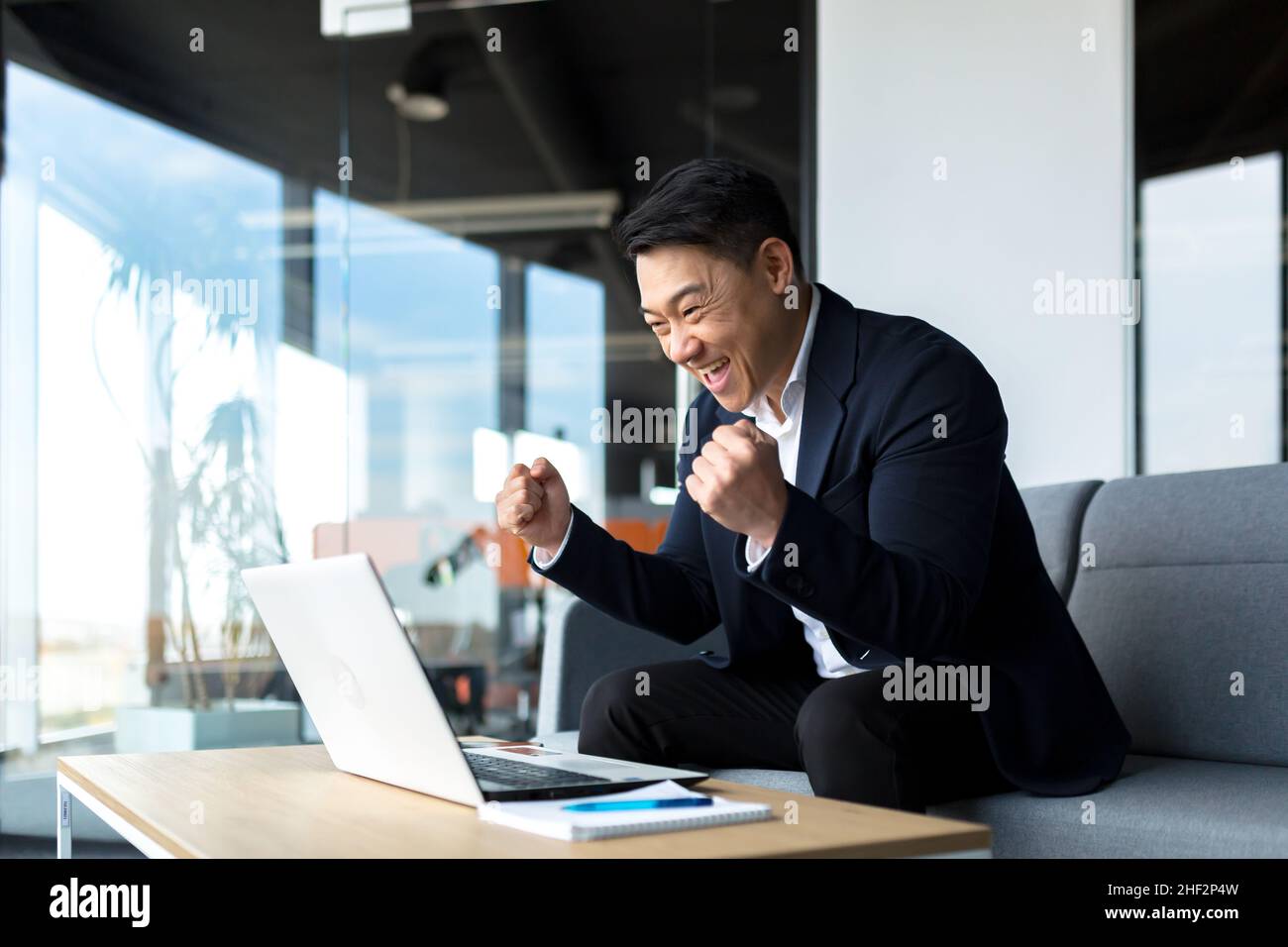 L'uomo d'affari ASIATICO gioisce nel successo, guarda lo schermo del portatile felice e sorride, ha ottenuto un grande risultato del suo lavoro, celebra la vittoria seduta in Foto Stock