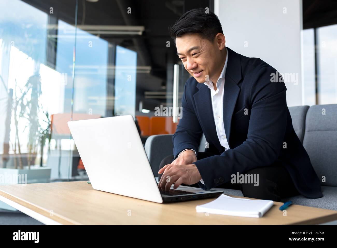 Uomo asiatico riuscito e felice che lavora in linea in remoto sul laptop sorridendo felice con il programma e l'operazione Foto Stock