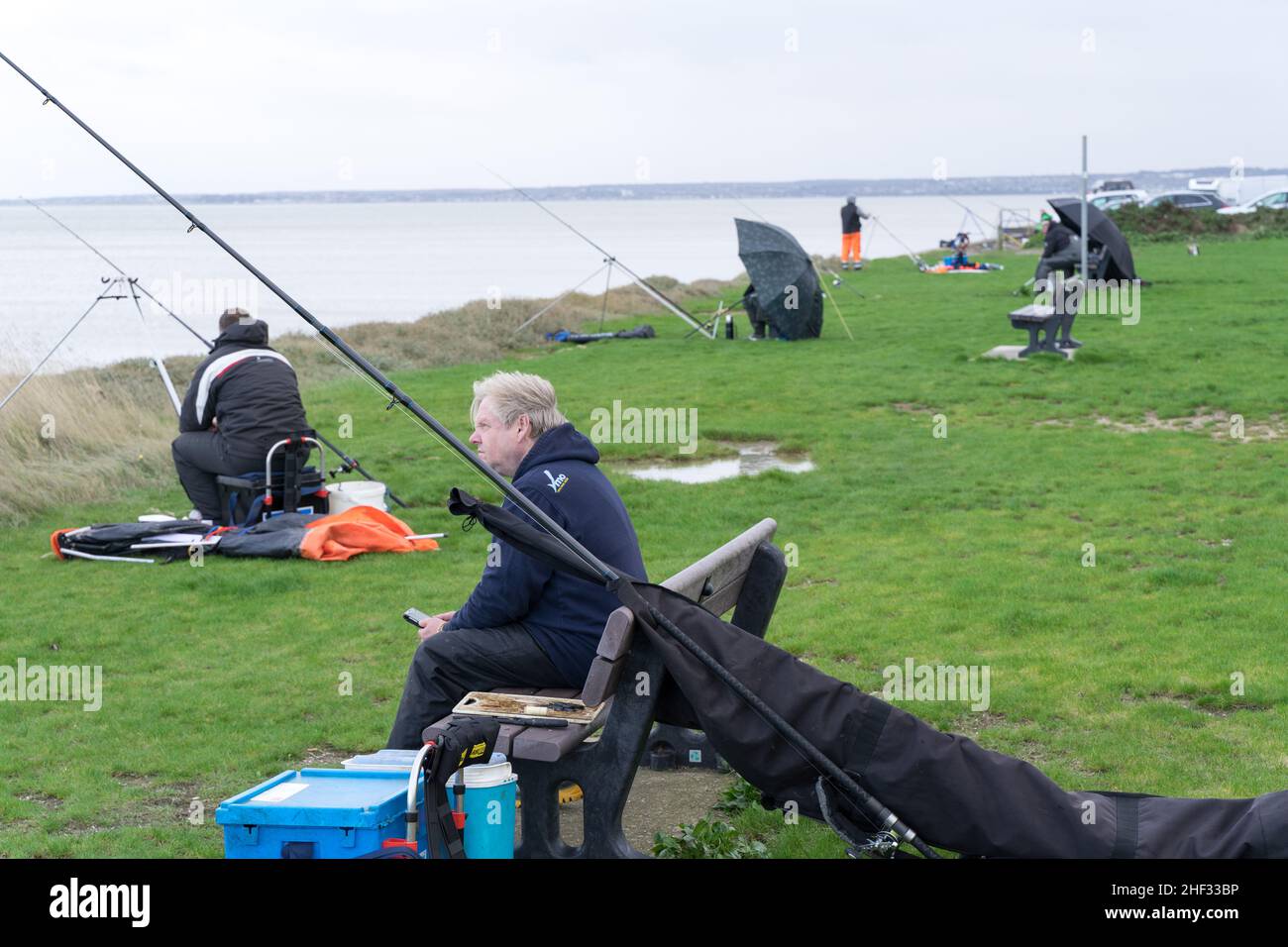 Un pescatore o un pescatore si siede sulla panchina della costa di Sheerness sulla mattina bagnata ventosa con l'attrezzatura da pesca e la pesca dal suo fianco Kent Inghilterra UK Foto Stock