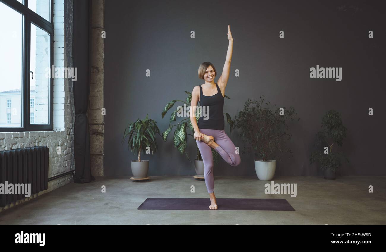 Donna solitaria in Sportswear fare fianco in uno spazioso studio Yoga. Sport Fit Woman Practices Hatha Yoga. Lunghezza intera. Sfondo grigio. Foto di alta qualità Foto Stock