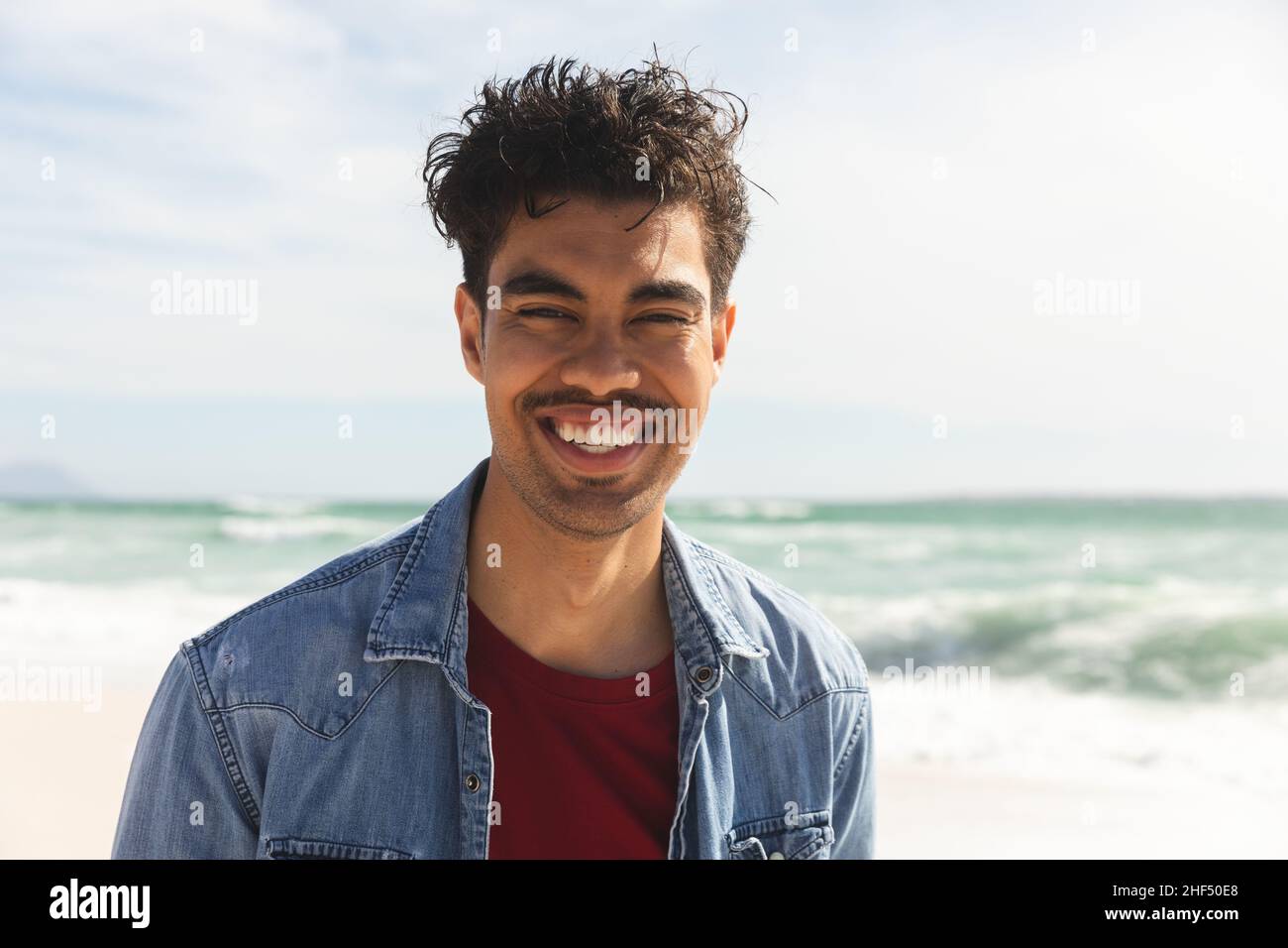 Ritratto di uomo biraciale sorridente che indossa una camicia in denim in spiaggia il giorno di sole Foto Stock
