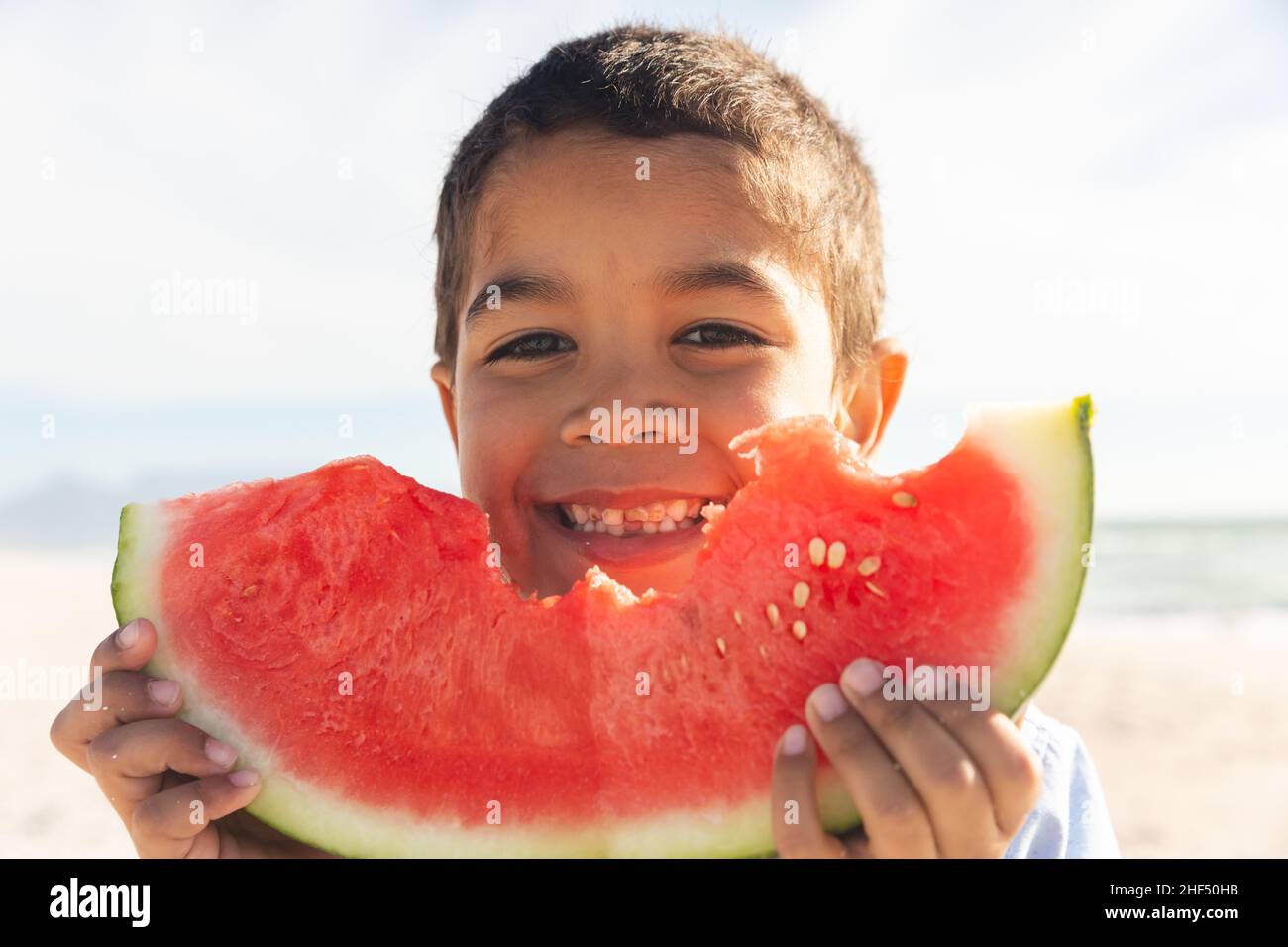Ritratto di sorridente ragazzo biraciale che tiene grande fetta di cocomero fresco in spiaggia il giorno di sole Foto Stock