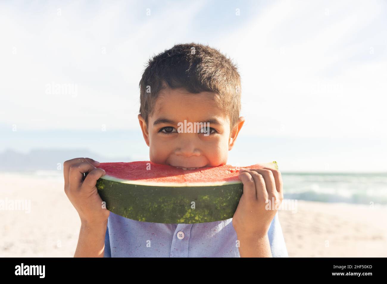 Ritratto di ragazzo biraciale carino mangiare grande fetta di cocomero fresco in spiaggia il giorno di sole Foto Stock