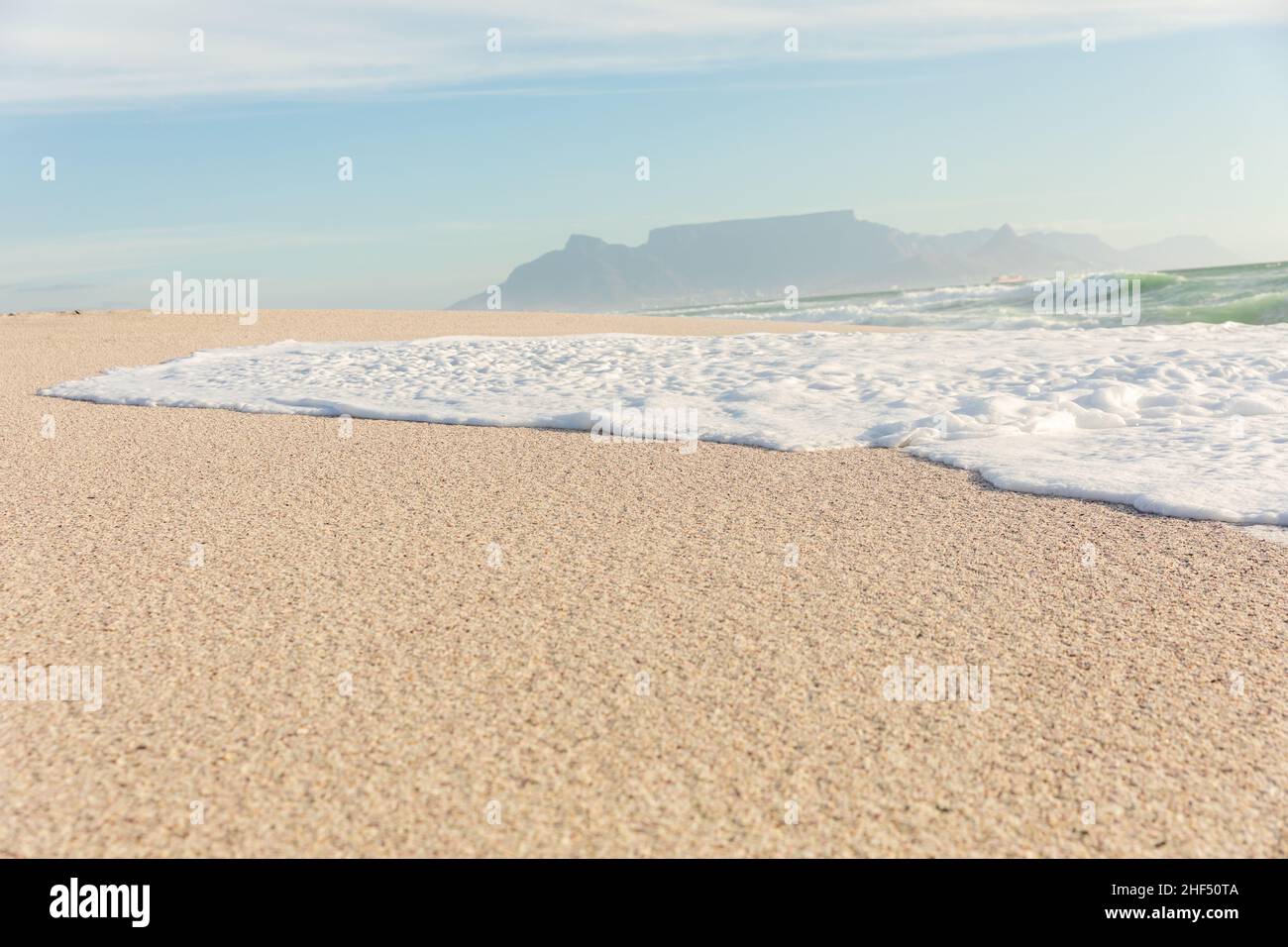 Schiuma d'onda di mare sulla riva a spiaggia soleggiata con montagne sullo sfondo distante Foto Stock