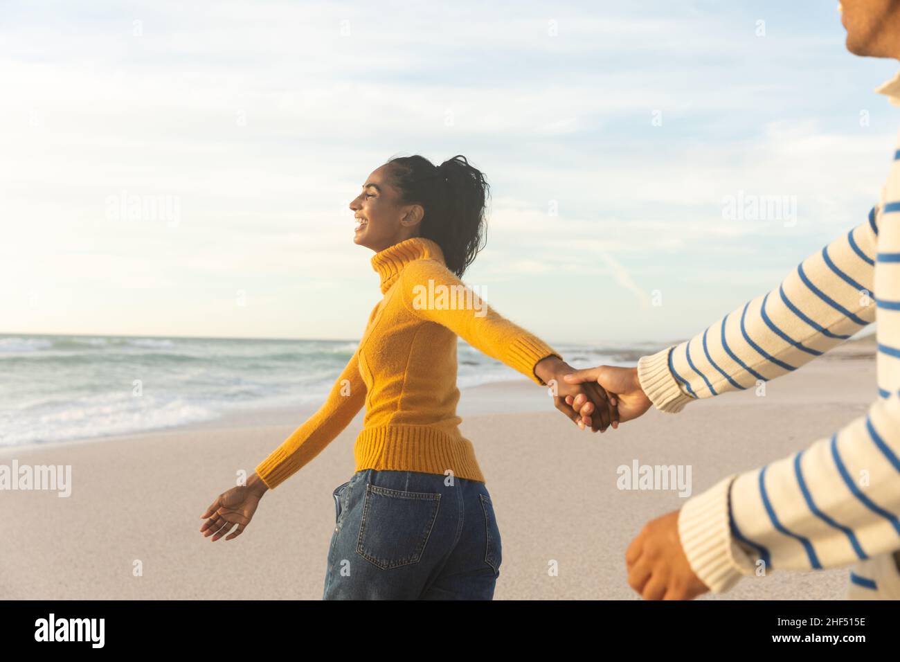 Allegra giovane donna biraciale che cammina mentre tiene una mano di ragazzo in spiaggia durante il tramonto Foto Stock