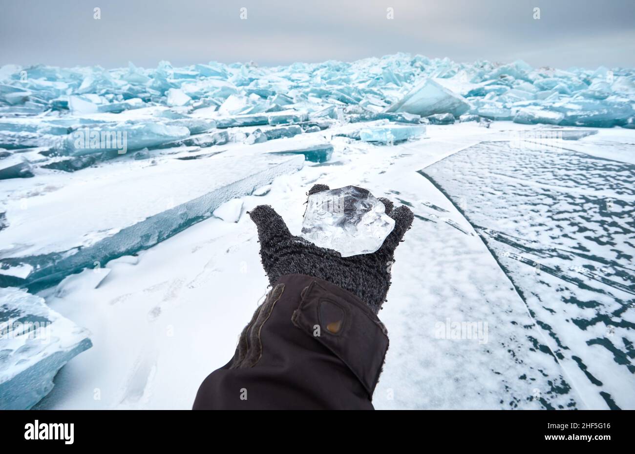 Aprire la mano nel guanto tenendo un piccolo pezzo trasparente di ghiaccio al lago ghiacciato con la neve Foto Stock