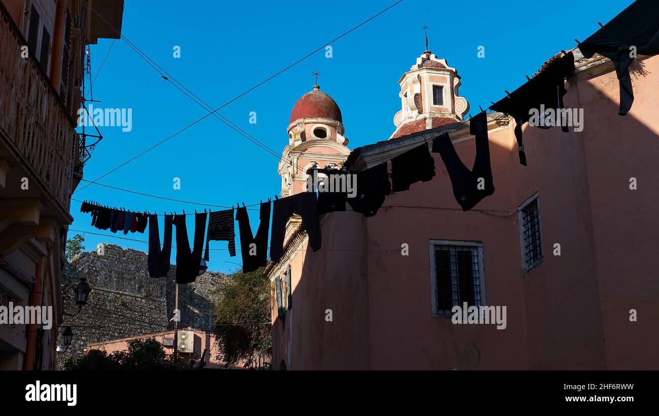 Grecia, Isole greche, Isole IONIE, Corfù, Corfù, Centro storico, vicolo della città vecchia all'ombra, in mezzo distanza clothesline con lavanderia all'ombra, nello sfondo chiesa con due torri chiesa rossa, cielo blu Foto Stock