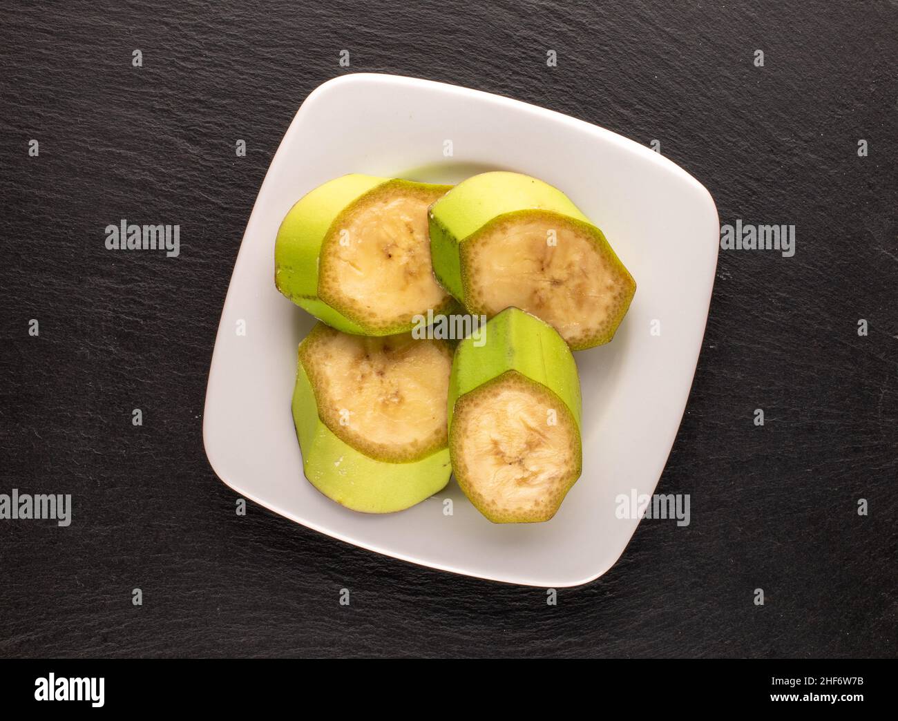 Quattro fette di una banana verde organica in un piatto bianco su una pietra ardesia, macro, vista dall'alto. Foto Stock