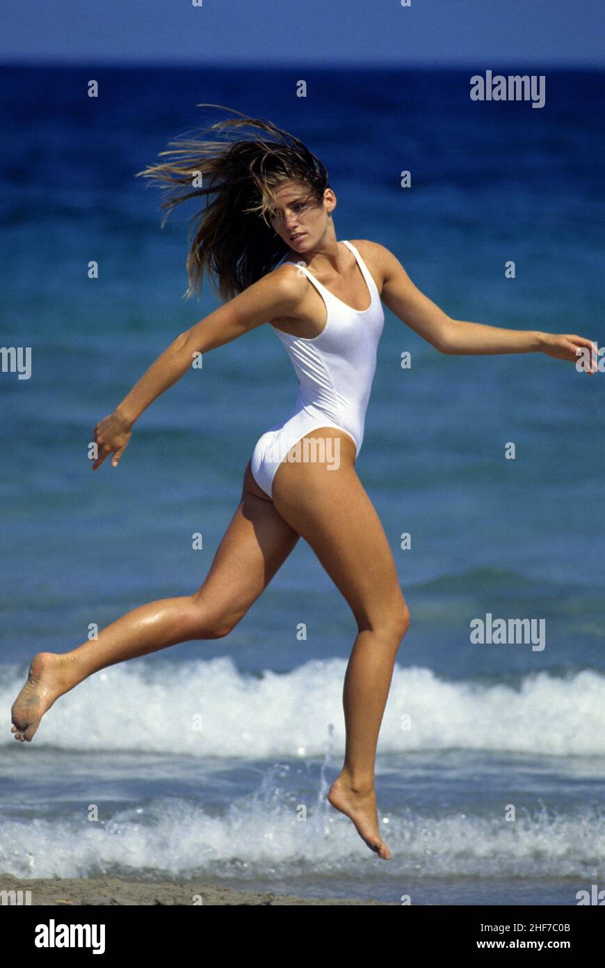 attraente biondo capelli giovane donna che corre sulla spiaggia blu mare sfondo naturale sorridente aspetto viso fotocamera anteriore bianco corpo tuta sportiva Foto Stock