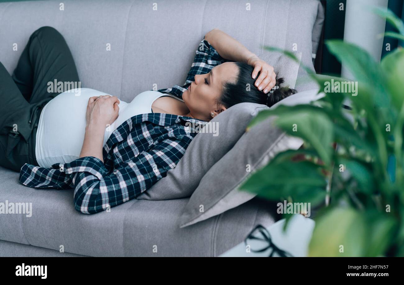la donna incinta riposa sul divano grigio e ricopre la testa con la mano nella disperazione a causa della depressione Foto Stock
