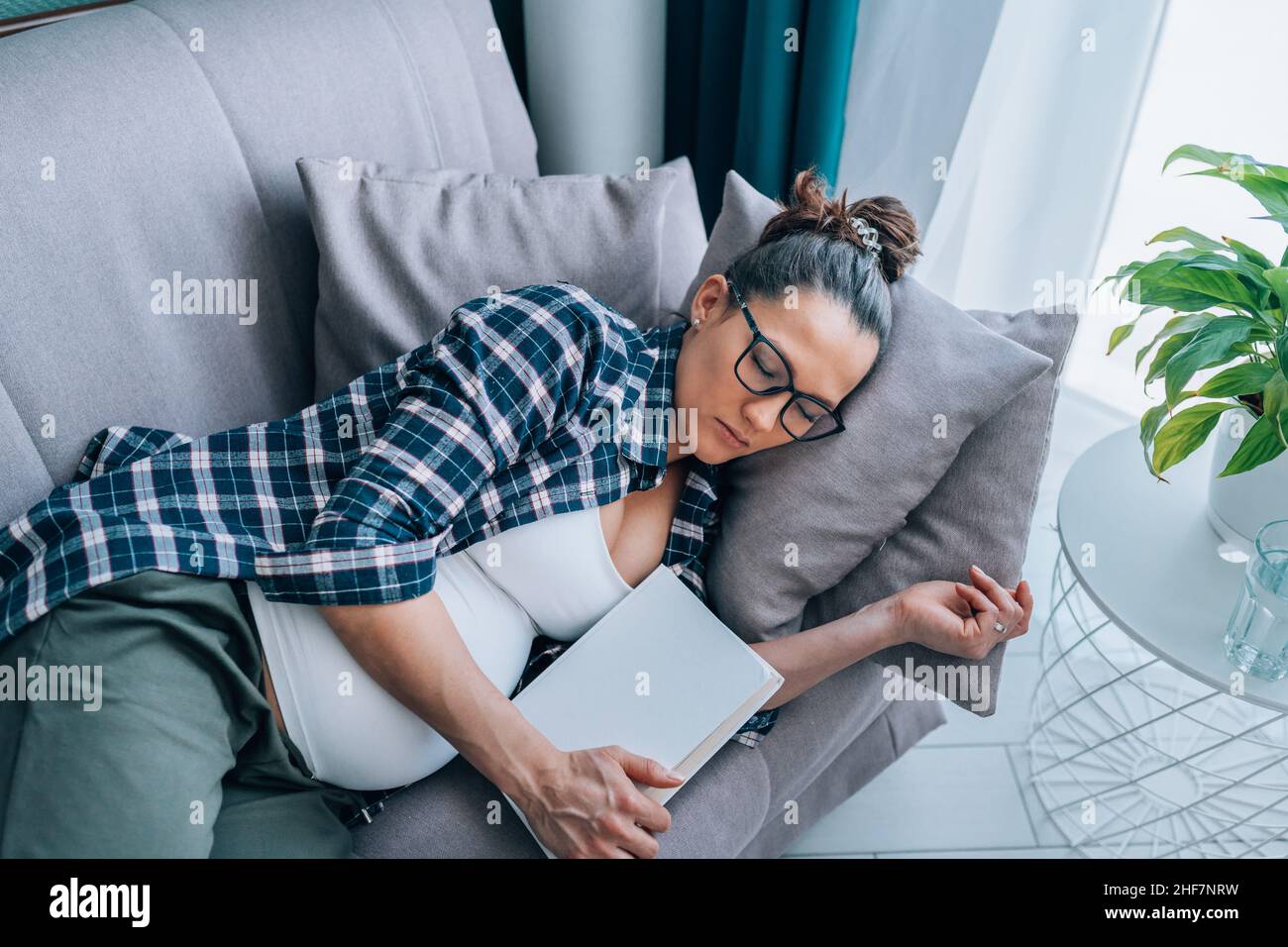 donna incinta in occhiali con camicia a scacchi dorme sdraiata sul divano grigio in soggiorno Foto Stock