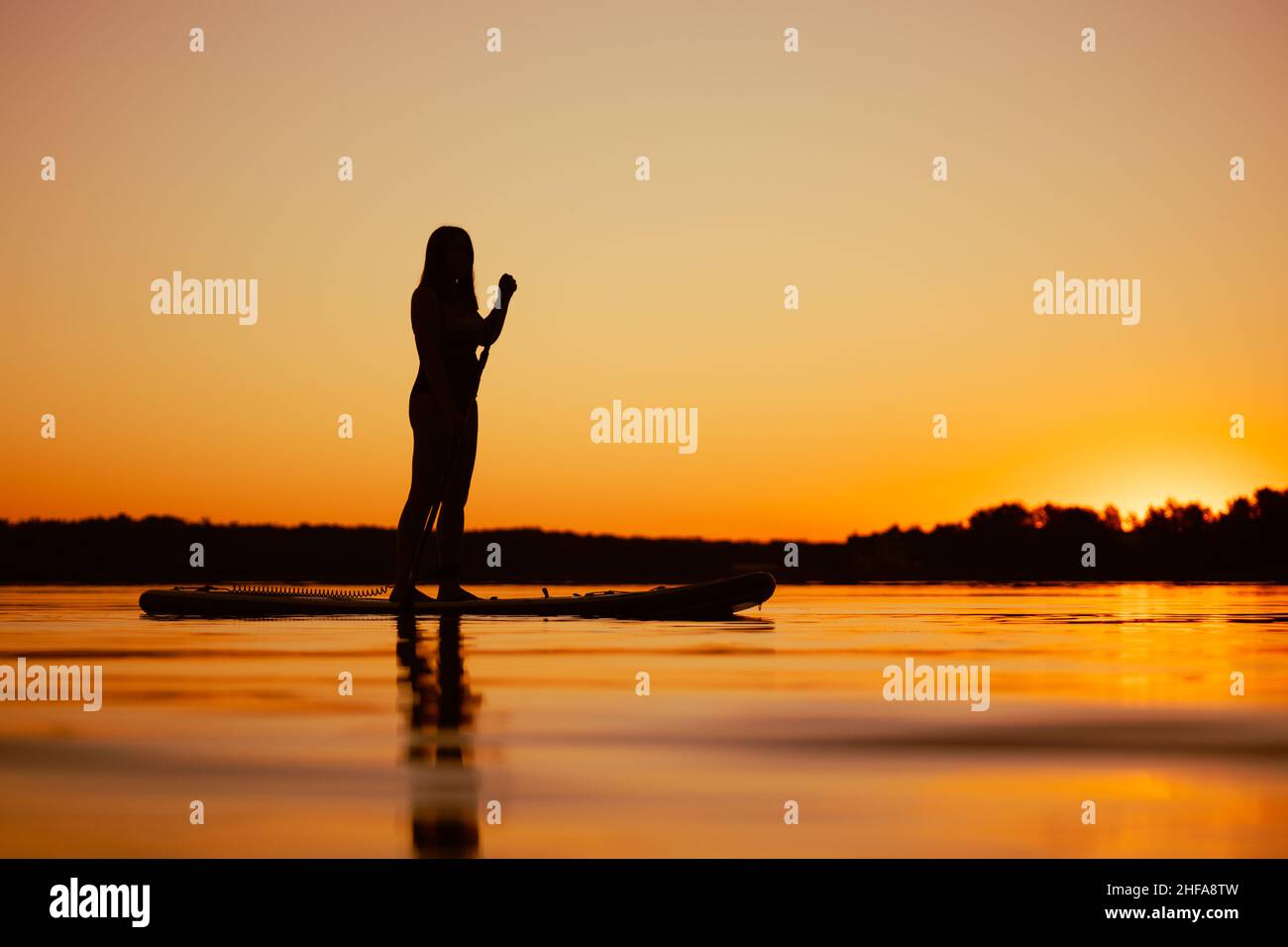 Donna scattata da un angolo basso sulla tavola sup con paddle in mano guardando il tramonto sorprendente che copre la superficie d'acqua di colore arancione in serata. Attivo Foto Stock