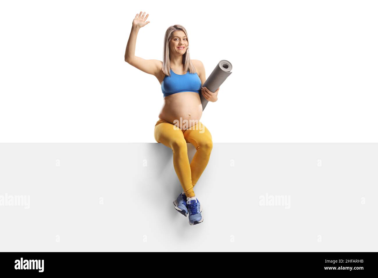 Donna incinta in abbigliamento sportivo con un tappeto da ginnastica seduto su un pannello vuoto e ondeggiante isolato su sfondo bianco Foto Stock