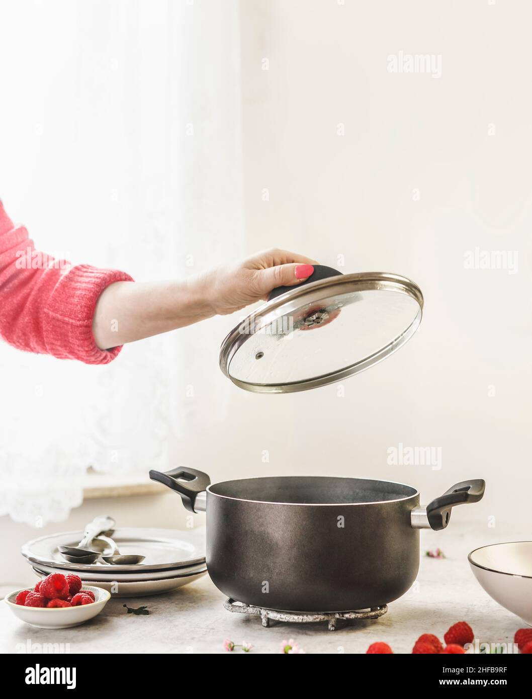 Mano donna in maglione rosso che tiene il coperchio della pentola sopra la pentola nera vuota di cottura sul tavolo con le stoviglie bianche e lamponi sullo sfondo della parete con l naturale Foto Stock