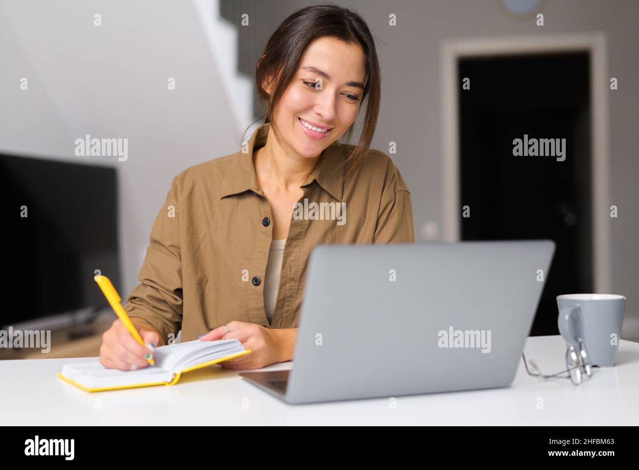 Sorridente giovane donna lavora da casa. Si siede al tavolo della cucina di fronte al portatile e notebook. Ragazza attraente Foto Stock