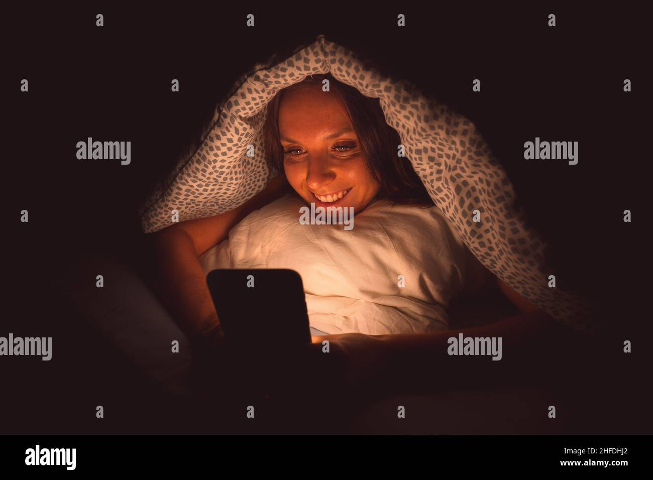 Donna sorridente felice sdraiata a letto sotto la coperta e digitando sullo smartphone la notte prima di dormire. Dipendenza dai social media e comunicazione Foto Stock