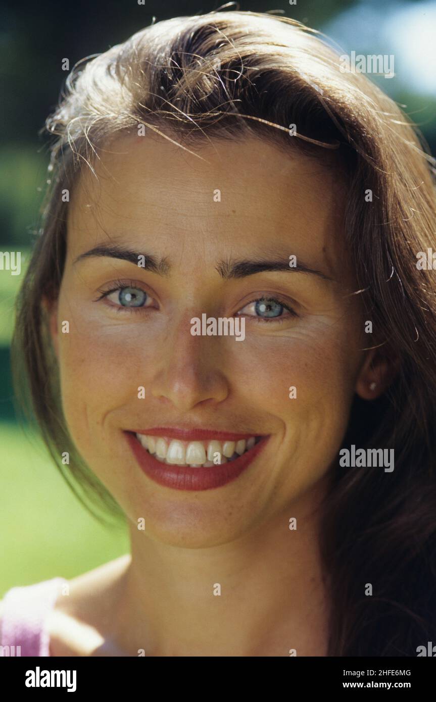 sorridente felice capelli scuri giovane donna primo piano ritratto guardando anteriore fotocamera verde fogliame sfondo blu occhi Foto Stock