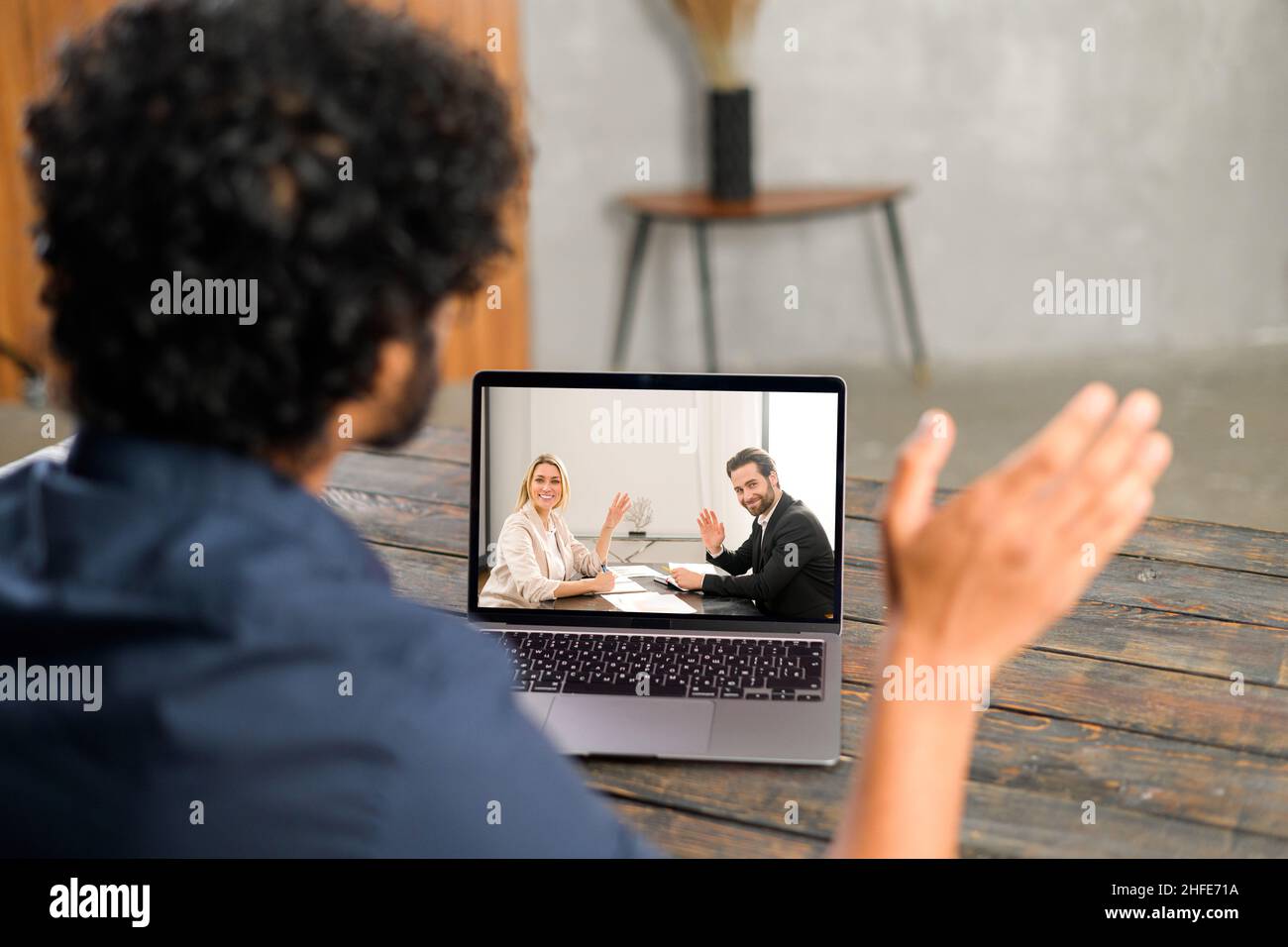 Vista in vita del giovane uomo in camicia blu che ondola con la mano e che dice ciao mentre chiacchiera con i colleghi di casa via computer portatile Foto Stock